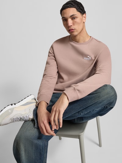 Męski model nosi Regular Fit Sweatshirt mit Logo-Stitching von PUMA PERFORMANCE z mieszanki bawełny, okrągły dekolt, elastyczne wykończenia, widoczne wyhaftowane logo, długość tyłu 69 cm, długość rękawów 69 cm, widoczne kolory beżowy i niebieski, struktura gładka, widok na cały korpus od przodu.
