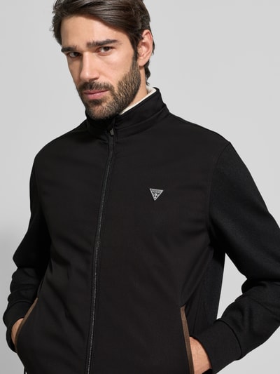 Männermodel trägt Windbreaker mit Zweiwege-Reißverschluss von Guess in Schwarz, Regular Fit, Label-Detail, Reißverschlusstaschen, Nahaufnahme Oberkörper.