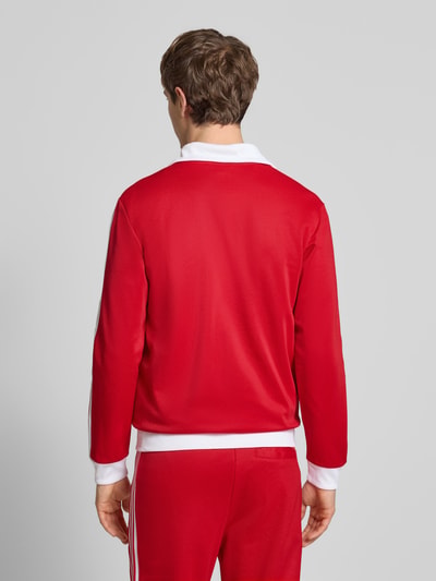 Männermodel trägt Regular Fit Sweatjacke mit Stehkragen Modell 'Classic' von adidas Originals in Rot mit weißen Details, Rückenansicht Ganzkörperansicht. Baumwoll-Mix, Logo-Stitching, Zweiwege-Reißverschluss, Eingrifftaschen mit Reißverschluss, elastische Abschlüsse, Rückenlänge 67 cm, Ärmellänge 64 cm.