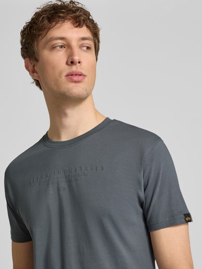 Męski model nosi T-Shirt aus Baumwolle von Alpha Industries w kolorze szarym, logo, prosty krój, okrągły dekolt, bawełna, długość tyłu w rozmiarze M: 68 cm, detale z logo, Ganzkörperansicht z przodu