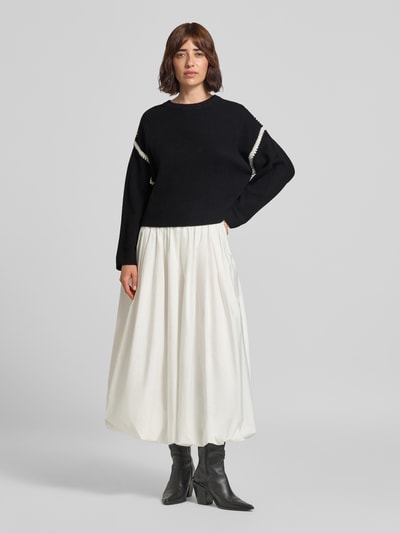 Frauenmodel trägt Strickpullover mit Rundhalsausschnitt und Kontraststreifen von Oui, Material-Mix mit Woll-Anteil, relaxed Fit, schwarzer Pullover mit weißen Akzenten, kombiniert mit weißem Rock und schwarzen Stiefeln, Ganzkörperansicht von vorne.