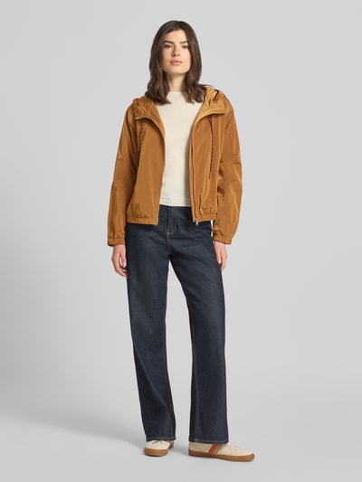 Kobiece model nosi Trekking Jacke mit Kapuze und Reißverschlusstaschen od Rich & Royal w kolorze karmelowym, prosty krój, kaptur z trokiem, zamek błyskawiczny, kieszenie zapinane na zamek błyskawiczny, elastyczne wykończenia, długość tyłu w rozmiarze 36: 58 cm, szerokość ramion w rozmiarze 36: 48 cm, szerokość klatki piersiowej w rozmiarze 36: 56 cm, długość rękawów w rozmiarze 36: 59 cm, gładka tekstura, widok całej sylwetki z przodu.