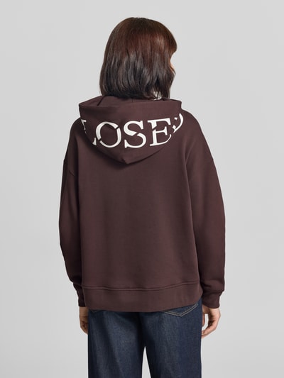 Frauenmodel trägt Oversized Hoodie aus Baumwolle von Closed in Dunkelbraun mit weißem Print, lockere Passform, Ganzkörperansicht Rückansicht.