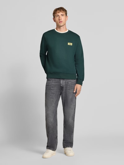 Männermodel trägt Sweatshirt mit Label-Print Modell 'RUSH' von JACK & JONES aus katoenmix in donkergroen met ribboorden, los model, ronde hals, ruglengte maat M 67 cm, mouwlengte maat M 67 cm, labelprint op de borst, vooraanzicht geheel lichaam.