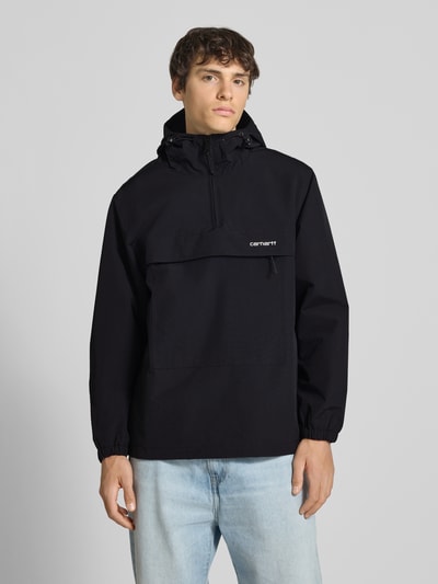 Männermodel trägt Windbreaker mit Label-Stitching von Carhartt Work In Progress in Schwarz, Regular-Fit, mit Kapuze, Reißverschluss und Tunnelzug, Rückenlänge 73 cm, Schulterbreite 48 cm, Brustweite 55 cm, Ärmellänge 66 cm, Ganzkörperansicht von vorne.