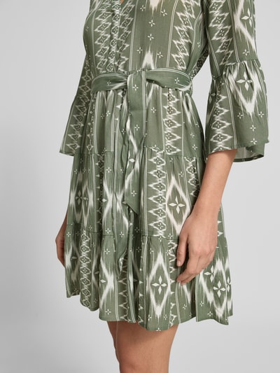 Frauenmodel trägt Tailliertes Tunikakleid aus reiner Viskose Modell 'MELBA' von Vero Moda, Optik:Allover-Muster, Mini-Länge, V-Ausschnitt, Bindegürtel, Trompetenärmel, Volant-Saum, Material: reine Viskose, Grüntöne mit weißem Muster, Textur weich, Nahaufnahme Oberkörper