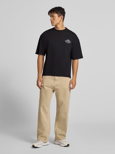Männermodel trägt Regular Fit T-Shirt aus reiner Baumwolle mit rückseitigem Print von REVIEW, schwarze Farbe, Logo-Detail, Ganzkörperansicht von vorne. Rückenlänge bei Größe M: 67 cm.