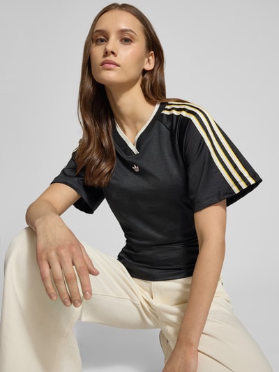 Vrouwenmodel draagt T-shirt met V-Ausschnitt van adidas Originals, zwart met labeldetails, asymmetrisch, aansluitende fit, korte mouw; close-up bovenlichaam.