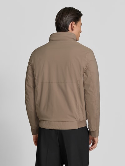 Männermodel trägt Regular Fit Jacke mit stójka von bugatti w kolorze beżowym, wpuszczane kieszenie, zamek błyskawiczny, elastyczny pas, długość tyłu w rozmiarze 50: 67 cm, szerokość ramion w rozmiarze 50: 45 cm, szerokość klatki piersiowej w rozmiarze 50: 58 cm, długość rękawów w rozmiarze 50: 67 cm, widok całej sylwetki z tyłu.