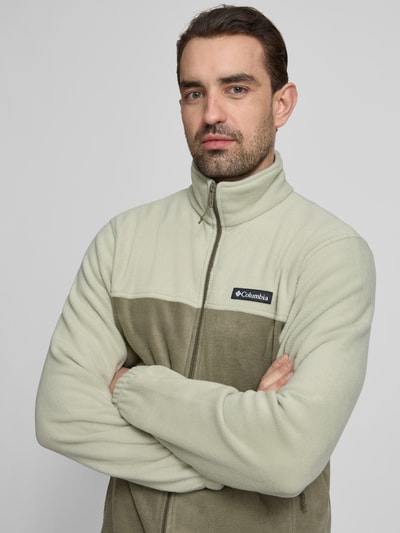 Männermodel trägt Regular Fit Fleecejacke mit Logo-Patch Modell 'Mountain' von COLUMBIA in Beige und Oliv, Nahaufnahme Oberkörper. Herren, Stehkragen, Reißverschluss, Eingrifftaschen, elastische Ärmelabschlüsse, Rückenlänge 70 cm, Ärmellänge 68 cm.