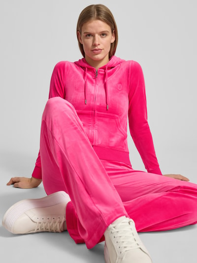 Frauenmodel trägt Sweatjacke mit Label-Stitching Modell 'ROBERTSON' von Juicy Couture im Pink, Material-Mix mit Stretch-Anteil, Regular Fit, Kapuze mit Tunnelzug, Reißverschluss und Känguru-Tasche, Ganzkörperansicht von vorne.
