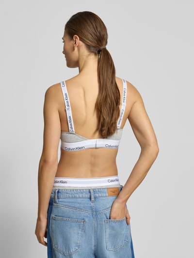 Kobiece model prezentuje Biustonosz typu bralette z pasem z logo i ramiączkami z logo od Calvin Klein Underwear, dopasowany krój, dekolt w serek, szara, gładka faktura, widok całej sylwetki z tyłu.