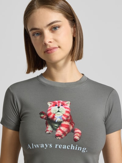 Modelka nosi Slim Fit T-Shirt mit Motiv-Print von DAISY STREET z mieszanki bawełny, okrągły dekolt, z krótkim rękawem, długość tyłu w rozmiarze S: 46 cm, szarość, motyw kolorowego kota, gładka tekstura, nadruk z napisem, widok na górną część ciała z przodu.