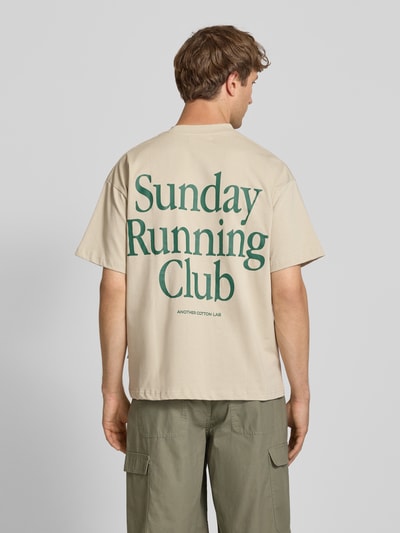 Męski model nosi T-Shirt mit Print und Rundhalsausschnitt von ANOTHER COTTON LAB z bawełny, krój relaxed fit, beżowy z zielonym nadrukiem, długość tyłu w rozmiarze M: 69 cm, całkowity widok z tyłu.