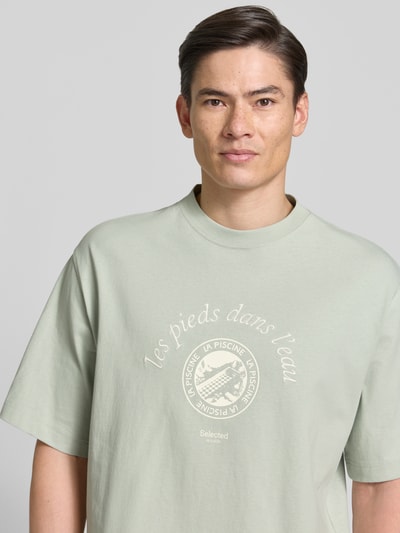 Männermodel trägt Relaxed Fit T-Shirt aus reiner Baumwolle Modell 'BOXYNORMAN GRAPHIC' von SELECTED HOMME in hellem Grün, Motiv-Print, Nahaufnahme Oberkörper.