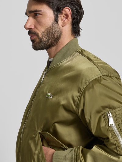 Mannelijk model draagt Regular Fit Bomberjack uit satijn van Lacoste in effen groen, waterafstotend, logopatch, zij- en napoleonzakken, warmteisolerend, geribde boorden, collegekraag, ritssluiting. Nahaufnahme bovenlichaam.