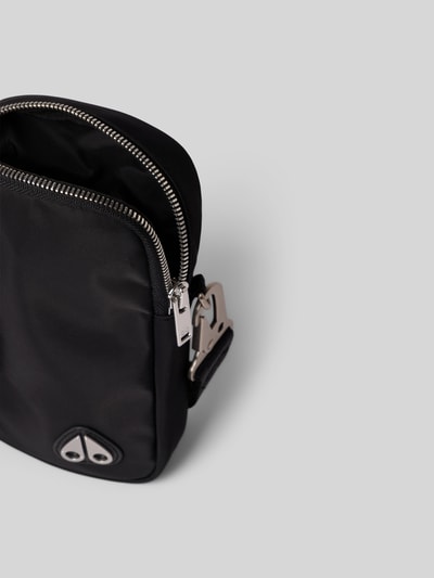 Produktfoto, Crossbody-Bag mit Logo-Detail von Moose Knuckles in Schwarz mit Zweiwege-Reißverschluss, Hauptfach, abnehmbarer Tragehenkel, verstellbarer Trageriemen, Maße ca. 11 cm Breite, 18 cm Länge, 4 cm Tiefe, glatte Struktur, silberfarbene Metalldetails.