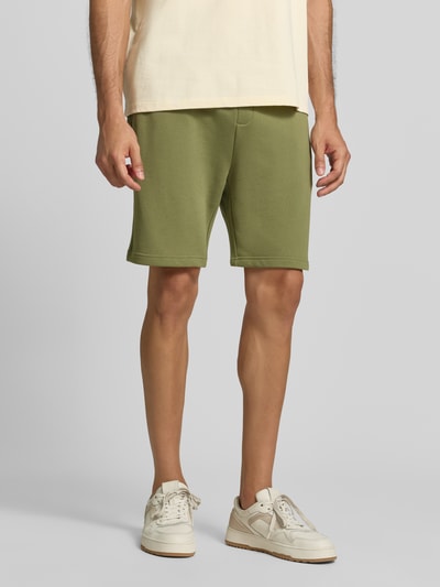 Männermodel trägt Shorts mit Label-Badge und elastischem Bund von Scotch & Soda in Olive, reine Baumwolle, Regular Fit, Ganzkörperansicht von vorne.
