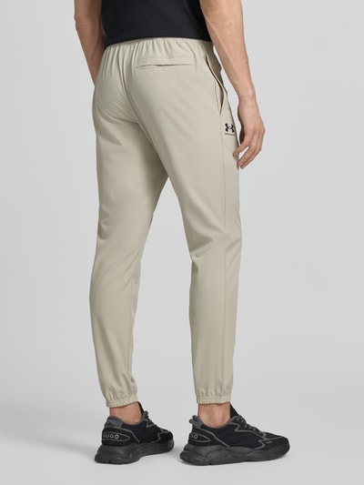 Mannenmodel draagt Regular fit jogger met logo-print model 'Vibe' van Under Armour in beige met elastische tailleband, zijzakken en elastische zoom, elastische materialenmix, productcontext uit beschrijving, volledige achteraanzicht.