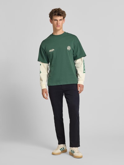 Męski model nosi Straight Fit Chino mit Eingrifftaschen Modell 'Vance' od Jack & Jones, ciemny materiał, Ganzkörperansicht von vorne. Chinos z elastycznego bawełnianego materiału, krój straight fit, kieszenie boczne i tylne, zapięcie na guzik i zamek, szerokość w rozmiarze 32/32: 43 cm, długość nogawki wewnętrznej w rozmiarze 32/32: 67 cm.