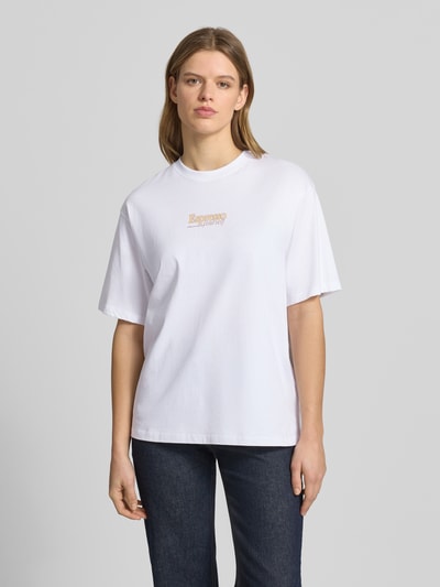 Kobiece modelka nosi damski t-shirt z bawełny model 'LATTE' od JJXX, krój loose fit, okrągły dekolt, długość tyłu w rozmiarze S: 65 cm, jasnobiały kolor, gładka tekstura, przód z napisem, całopostaciowy widok z przodu.