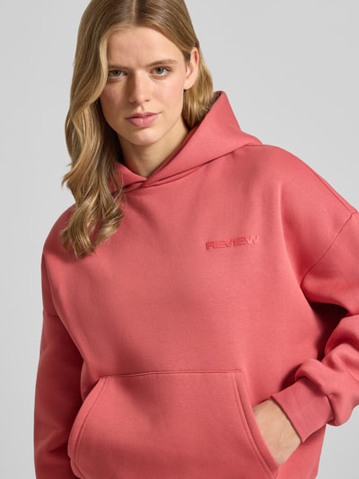 Frauenmodel trägt Hoodie mit Logo und Känguru-Tasche von REVIEW in Rosa, Baumwoll-Mix, Relaxed-Fit, Nahaufnahme Oberkörper.