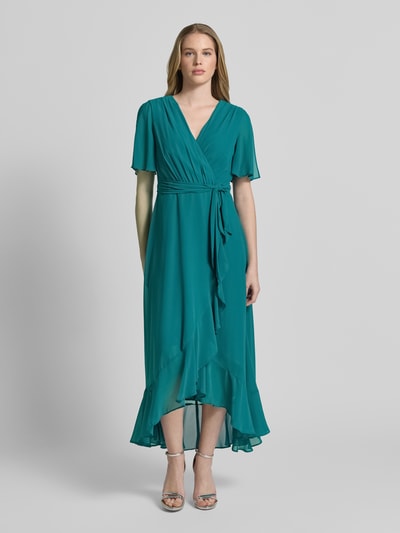 Frauenmodel trägt Cocktailkleid in Wickel-Optik mit Volantsaum von Christian Berg Cocktail in Türkis, Chiffon, tailliert, Midikleid, V-Ausschnitt, Schmetterlingsärmel, Bindegürtel, blickdichtes Unterkleid, Ganzkörperansicht von vorne.