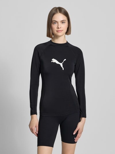 Frauenmodel trägt Badeshirt im langärmeligen Design Modell 'RASH GUARD' von PUMA, mieszanka materiałów z dodatkiem streczu, prosty krój, okrągły dekolt, nadruk z logo, uv-protection 50+, odporne na chlor, długość tyłu w rozmiarze S: 56 cm, czarny kolor, gładka faktura, Ganzkörperansicht von vorne.