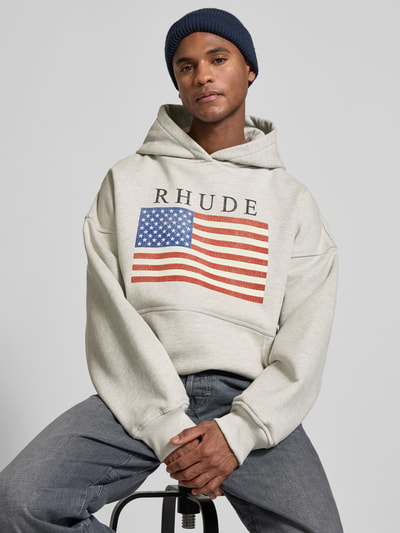 Männermodel trägt Oversized Hoodie mit Logo-Print von RHUDE aus reiner Baumwolle, hellgrau mit Flaggenmotiv, Nahaufnahme Oberkörper.
