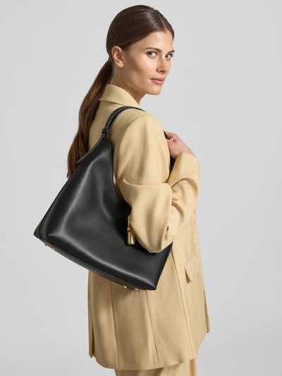 Frauenmodel trägt Tote Bag mit Logo-Detail Modell 'Goccia' von FURLA in Schwarz, Hauptfach mit Riemenverschluss, Schmale Trageriemen, Breite 33 cm, Länge 31 cm, Tiefe 12 cm, Schulterriemen 51 cm, Ganzkörperansicht Rückansicht.