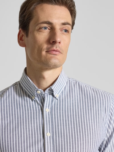 Męski model nosi Relaxed Fit Freizeithemd mit Button-Down-Kragen von Lindbergh, mieszanka materiałów z dodatkiem streczu, wzór w paski, listwa guzikowa, długość tyłu w rozmiarze M: 76 cm, szerokość w rozmiarze M: 44 cm, długość rękawów w rozmiarze M: 64 cm, jasne i niebieskie pionowe paski, struktura gładka, zbliżenie na górną część ciała