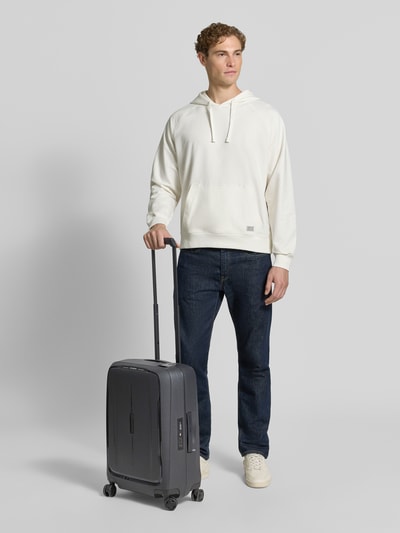 Männermodel trägt Koffer mit Strukturmuster Modell 'Spinner' von Samsonite, Hardshell in Grau, Hauptfach mit Reißverschluss, Ganzkörperansicht von vorne.