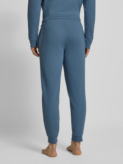 Męski model nosi Relaxed Fit Pyjama-Hose aus Baumwoll-Mix Modell 'AUSTIN PANTS' od HUGO, kolor niebieski, faktura waflowa, regular fit, kieszenie boczne, nadruk z logo, zdjęcie całej sylwetki od tyłu.