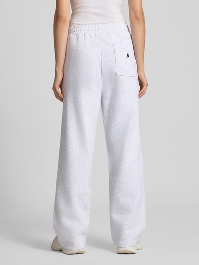 Kobiece modelka nosi spodnie dresowe Wide Fit Sweatpants aus reiner Baumwolle Model 'Cassie' od Carhartt Work In Progress, jasnoszare, wyhaftowane logo, elastyczny pas, bawełna, długość wewnętrzna nogawki 76 cm w rozmiarze S, widok całej sylwetki z tyłu.