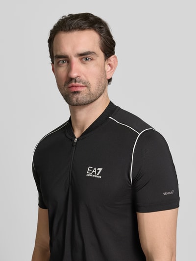 Männermodel trägt Regular Fit T-Shirt mit Stehkragen von EA7 Emporio Armani in Schwarz mit Reißverschluss und Label-Print, Nahaufnahme Oberkörper.