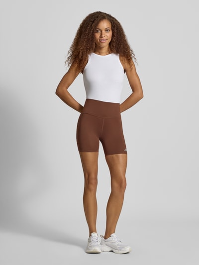 Frauenmodel trägt High Rise Radlershorts mit Logo-Print Modell 'HARMONY' von New Balance in Braun, körpernaher Schnitt, elastischer Bund, Ganzkörperansicht von vorne.