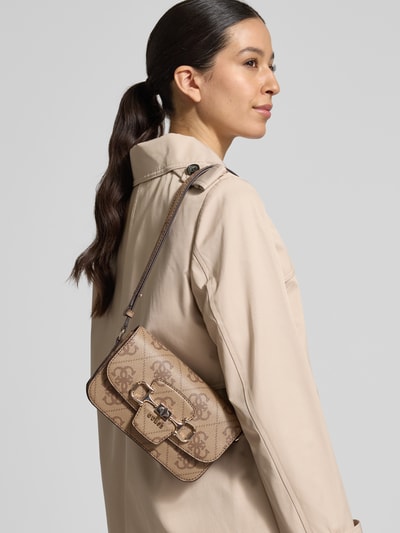 Modelka kobieca nosi torbę na ramię Umhängetasche mit Label-Detail Modell 'JANIE' od Guess, beżowy kolor, detal z logo, pasek na ramię regulowane, główna przegroda z zapięciem na zatrzask, przegródka zapinana na zamek błyskawiczny, szerokość w rozmiarze 1: 21 cm, długość w rozmiarze 1: 14 cm, głębokość w rozmiarze 1: 5 cm, długość paska: 78 cm, tekstura skóry imitowanej, widok całej sylwetki z przodu.