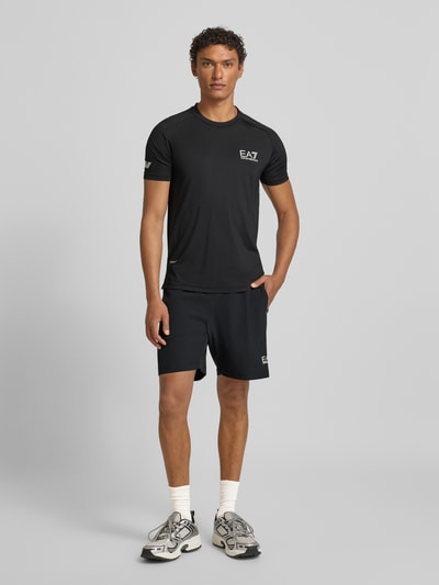 Männermodel trägt T-Shirt mit Label-Print von EA7 Emporio Armani, schwarzer Material-Mix mit Stretch-Anteil, Slim Fit, Ganzkörperansicht von vorne.
