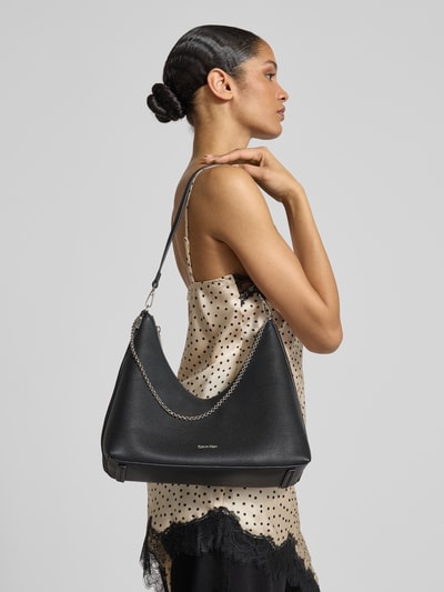 Kobiece modelka nosi torebkę Handtasche mit Label-Detail od CK Calvin Klein, czarny kolor, detal z logo, pasek na ramię, główna przegroda z zamkiem błyskawicznym, przegródka wewnętrzna zapinana na zamek błyskawiczny, szerokość w rozmiarze 1: 24 cm, długość w rozmiarze 1: 27 cm, głębokość w rozmiarze 1: 11 cm, długość paska na ramię w rozmiarze 1: 68 cm, gładka faktura, całkowity widok z boku