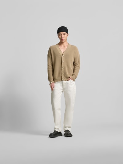 Männermodel trägt Regular Fit Cardigan im Used-Look von MM6 Maison Margiela in Beige, kombiniert mit weißer Hose, Ganzkörperansicht von vorne. Reine Baumwolle, Knopfleiste, Logo-Detail, lockerer Schnitt.