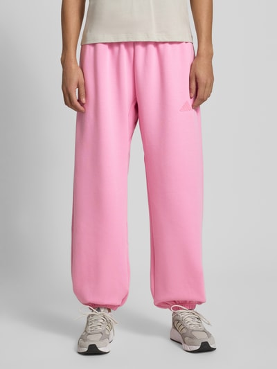 Frauenmodel trägt Loose Fit Sweatpants mit Baumwoll-Anteil von adidas Sportswear, Optik: Unifarben, Passform: Loose-Fit, Label-Patch, Bundabschluss: Elastisch mit Kordelzug, Beinabschluss: Elastisch, gerippt, seitliche Eingrifftaschen, Materialverarbeitung: Sweat, Farbe Pink, glatte Textur, Ganzkörperansicht von vorne.