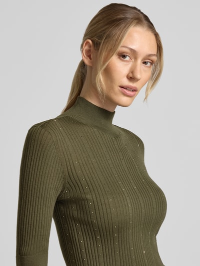 Frauenmodel trägt Slim Fit Longsleeve aus reiner Lanawolle Modell 'Maglia Costine' von Liu Jo White in Olivgrün, Stehkragen, 1/2-Arm, gerippte und semitransparente Textur mit dezenten Glanzakzenten, Nahaufnahme Oberkörper.