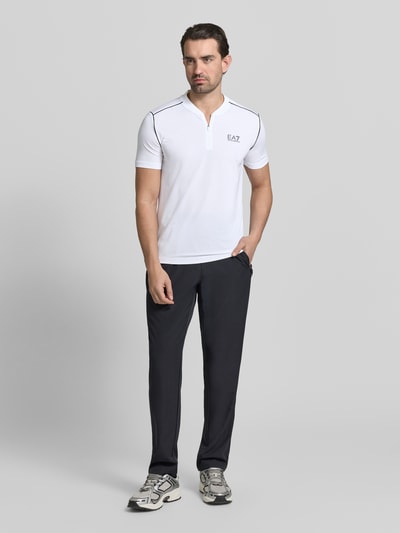 Männermodel trägt Regular Fit T-Shirt mit Stehkragen von EA7 Emporio Armani in Weiß, Reißverschluss, Label-Print, Ganzkörperansicht von vorne.