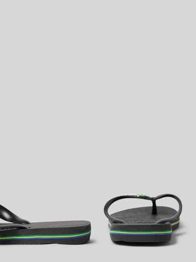 Produktfoto, schwarze Slippers mit Logo-Detail Modell 'Brasil' von Havaianas, offene Ferse, dezente grüne und gelbe Linien, rau strukturierte Sohle, abgerundete Passform, flacher Absatz, seitliche Ansicht. Damen Slippers, Absatzhöhe 2 cm, Schaftweite 8 cm.