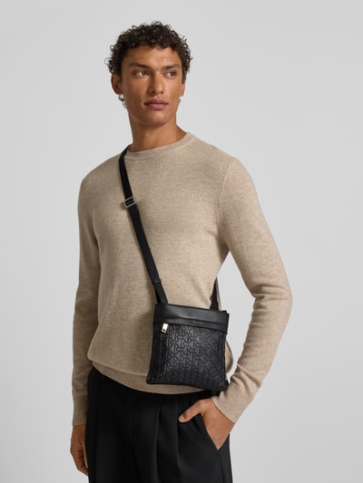 Männermodel trägt schwarze Handtasche mit Label-Detail und Schulterriemen von CK Calvin Klein, Allover-Look, Hauptfach mit Reißverschluss, Ganzkörperansicht von vorne.
