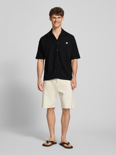 Männermodel trägt Regular Fit Freizeithemd mit Label-Patch Modell 'LIBCO' von Pegador, Baumwoll-Anteil, Reverskragen, schwarz, Knopfleiste, kurzärmelig, strukturierte Oberfläche; Kombination mit heller Shorts, Ganzkörperansicht von vorne.