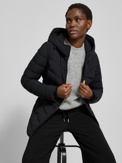 Frauenmodel trägt Daunenjacke mit Kapuze von Moose Knuckles in Schwarz, Material-Mix mit Stretch-Anteil, Taillierter Schnitt, Reißverschluss, Reißverschlusstaschen, Rückenlänge 75 cm, Brustweite 48 cm, Ärmellänge 65 cm, Ganzkörperansicht von vorne.