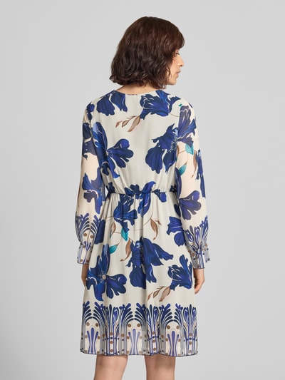 Frauenmodel trägt knielanges Kleid im Allover-Look Modell 'Ja44milaa' von ZABAIONE mit blauem Blütenmuster auf cremefarbenem Chiffon, A-Linie, V-Ausschnitt, Wickel-Optik, Rückenlänge 94 cm, Brustweite 48 cm, Ärmellänge 60 cm, Ganzkörperansicht Rückansicht.
