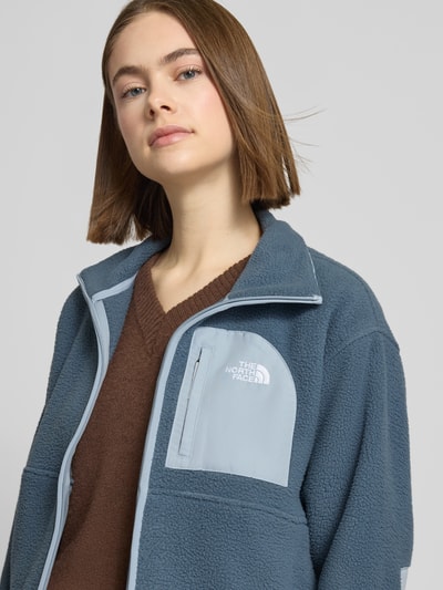 Modelka nosi Fleecejacke mit Stehkragen und Label-Stitching od The North Face, kolor niebieski, tekstura polarowa, krój regular fit, kieszeń na piersi, długość tyłu w rozmiarze S 57 cm, szerokość ramion w rozmiarze S 50 cm, szerokość w rozmiarze S 56 cm, długość rękawa w rozmiarze S 56 cm, zamek błyskawiczny, ujęcie: zbliżenie górnej części ciała z przodu.