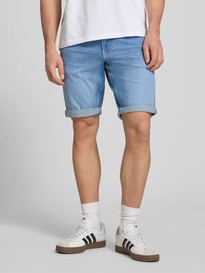 Männermodel trägt Regular Fit Jeansshorts aus Baumwoll-Mix Modell 'RONNIE SHORT' von Tommy Jeans in Denim-Optik mit Label-Patch, Logo-Stitching, 5-Pocket-Design, Gürtelschlaufen und Knopf-Verschluss, Farbton Hellblau, Materialstruktur glatt, Ganzkörperansicht von vorne.